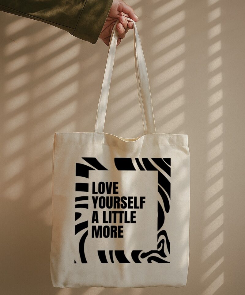 Tote bag with bold black typography “LOVE YOURSELF A LITTLE MORE” surrounded by abstract graphic lines.